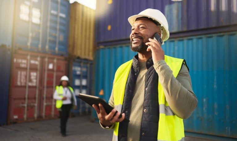 Logistics, shipping and construction worker on the phone with tablet in shipyard. Transportation engineer on smartphone in delivery, freight and international distribution business in container yard